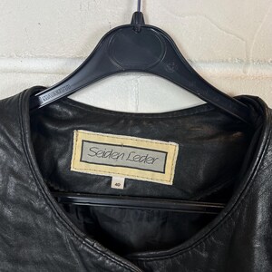 Op de afbeelding: Zwarte leren jas met een ronde hals, hangend aan een zwarte hanger. Het label leest "Seiden Leder" en maat 40. De jas is gemaakt van glad, glanzend leer.