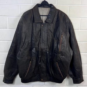 Könnte beinhalten: Eine dunkelbraune Lederjacke mit klassischem Kragen, einem durch eine Druckknopfleiste verdeckten Frontreißverschluss und zwei Reißverschlusstaschen an der Taille. Eine zusätzliche vertikale Reißverschlusstasche befindet sich auf der linken Brust.