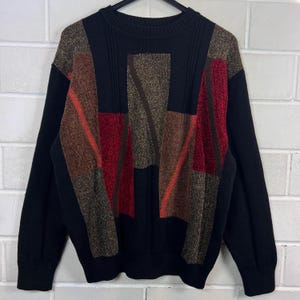 Könnte beinhalten: Schwarzer Pullover mit einem geometrischen Design in Braun-, Rot- und Goldtönen. Der Pullover hat einen Rundhalsausschnitt und lange Ärmel. Das Design besteht aus rechteckigen und diagonalen Formen.