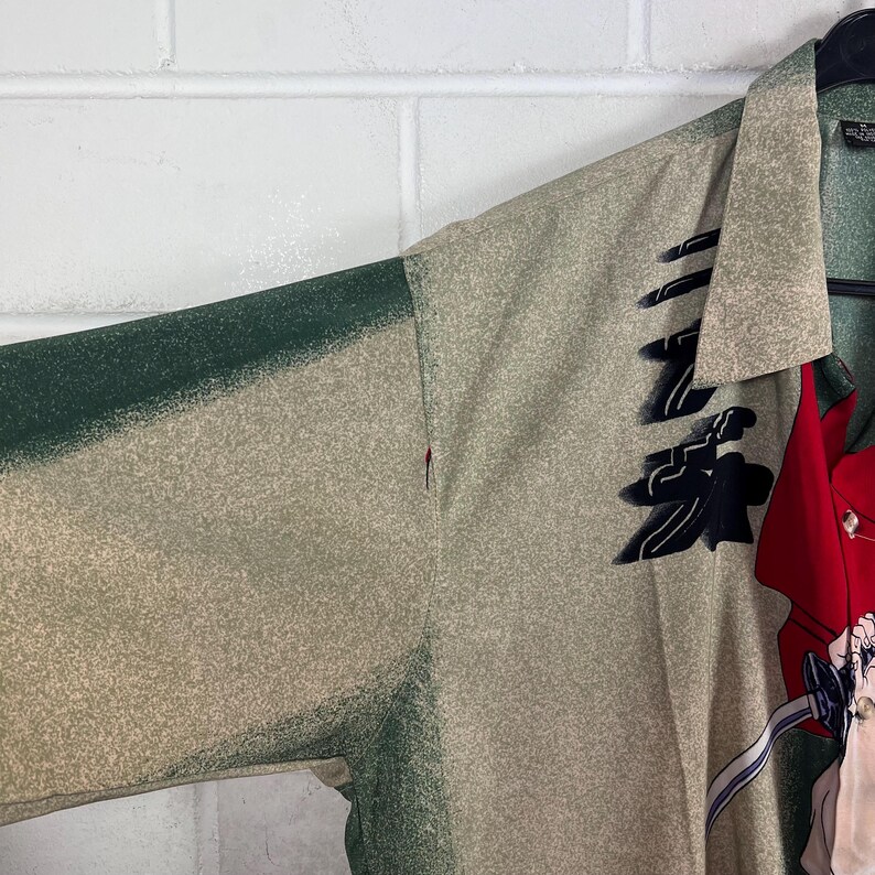 K&ouml;nnte beinhalten: Ein Button-Down-Hemd mit beigem und gr&uuml;nem Muster. Das Hemd hat einen gr&uuml;nen &Auml;rmel und ein rotes Panel mit einem grafischen Design. Es h&auml;ngt an einem schwarzen Kleiderb&uuml;gel vor einer wei&szlig;en Backsteinmauer.