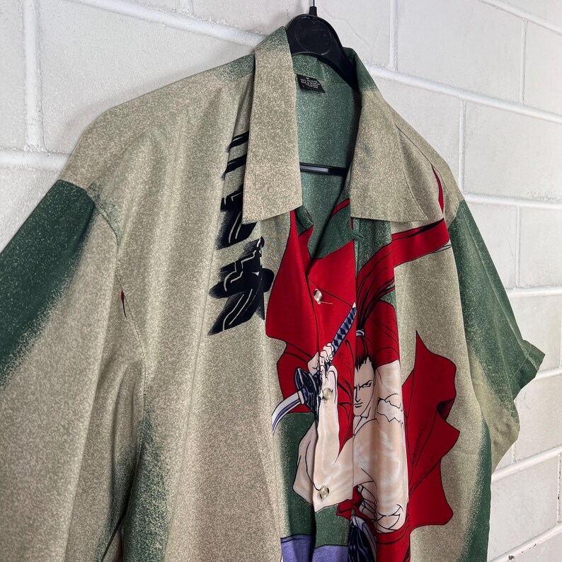 K&ouml;nnte beinhalten: Kurz&auml;rmeliges Button-Down-Hemd in Beige, Gr&uuml;n und Rot. Das Hemd zeigt eine Grafik eines Samurai-Kriegers mit Schwert und japanischen Schriftzeichen. Es h&auml;ngt an einem schwarzen Kleiderb&uuml;gel vor einer wei&szlig;en Backsteinmauer. Klassischer Kragen.