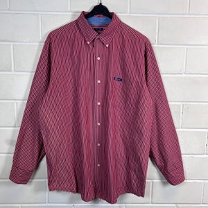 Könnte beinhalten: Ein langärmeliges Hemd mit Knopfleiste und vertikalem Streifenmuster in Rot, Weiß und Marineblau. Das Hemd verfügt über einen Button-Down-Kragen, eine Brusttasche mit einem kleinen Chaps-Markenetikett und weiße Knöpfe.