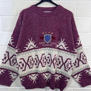Könnte beinhalten: Ein weinroter und weißer Strickpullover mit Rundhalsausschnitt. Der Pullover hat ein geometrisches Muster in Weiß und Grau mit einem Schildemblem mit der Aufschrift "Snow Team Boarding". Der Pullover hängt an einem schwarzen Kleiderbügel.