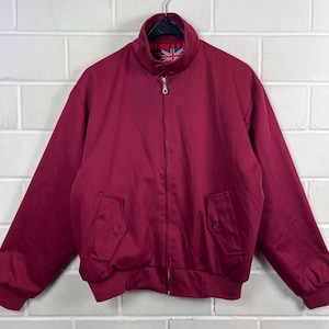 Può includere: Giacca bordeaux con zip frontale, due tasche anteriori e orlo e polsini a coste. La giacca ha un collo alto e un motivo Union Jack sulla fodera interna. La giacca è appesa a una gruccia nera.