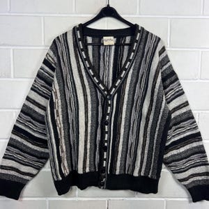 Könnte beinhalten: Ein Vintage-Cardigan mit Knopfleiste und vertikalem Streifenmuster in Schwarz-, Grau- und Beigetönen. Der Pullover hat einen V-Ausschnitt, lange Ärmel und schwarze Bündchen und Saum. Das Etikett lautet "Eagle's Eye".