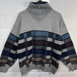 May include: A gray sweater with a high collar and a quarter-zip closure. The sweater features a horizontal plaid design in shades of gray, blue, and brown across the chest. The sleeves and upper body are solid gray.