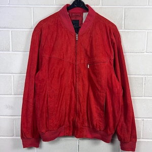 Peut inclure: Veste bomber en daim rouge vif avec fermeture éclair intégrale. Le blouson présente un col, des poignets et un ourlet côtelés, ainsi qu'une poche unique sur la poitrine. Suspendu à un cintre noir devant un mur de briques blanches.
