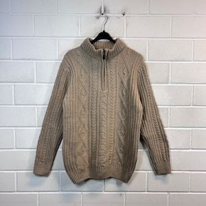 Peut inclure: Un pull en maille beige à câble avec une fermeture éclair à moitié. Le pull a un col et des poignets côtelés.