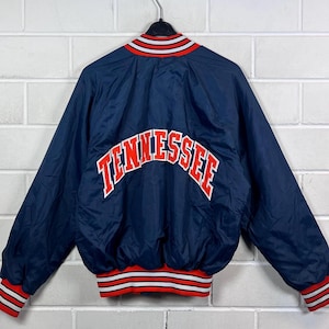 Könnte beinhalten: Eine marineblaue Bomberjacke mit dem Wort "TENNESSEE" in roter und weißer Schrift auf dem Rücken. Die Jacke hat einen rot-weiß-marineblauen Streifenbesatz an Kragen, Manschetten und Bund. Die Jacke hängt an einem schwarzen Kleiderbügel.