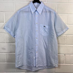 Könnte beinhalten: Hellblaues Kurzarmhemd mit Knopfleiste und Brusttasche. Das Hemd hat ein kleines grünes Krokodil-Logo auf der Tasche. Der Stoff wirkt leicht und eignet sich für warmes Wetter. Knöpfe sind sichtbar.