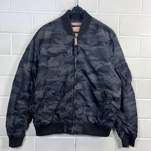 Könnte beinhalten: Eine schwarze Camouflage-Bomberjacke mit durchgehendem Reißverschluss, geripptem Kragen und Bündchen. Die Jacke hat eine kleine Tasche am linken Ärmel und zwei Vordertaschen. Die Jacke hängt an einem schwarzen Kleiderbügel.