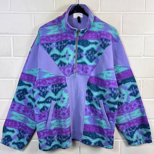 Könnte beinhalten: Ein Fleece-Pullover mit kurzem Reißverschluss und einem lebendigen geometrischen Muster in Violett, Türkis und Dunkelblau. Das Kleidungsstück verfügt über ein einfarbiges hellviolettes Mittelteil, einen hohen Kragen und zwei seitliche Taschen.