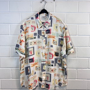 Peut inclure: Une chemise boutonnée à manches courtes blanche avec un imprimé abstrait coloré comportant des chiffres et des formes géométriques. La chemise a un col et une fermeture boutonnée.