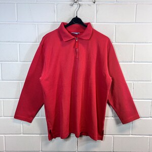 Può includere: Una camicia rossa a maniche lunghe con collo e cerniera a un quarto di lunghezza. La camicia è appesa a una gruccia nera contro un muro di mattoni bianchi. Le maniche sono lunghe e l'orlo inferiore ha spacchi laterali.