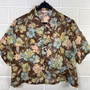 Könnte beinhalten: Ein kurzärmeliges, durchgeknöpftes Hemd aus transparentem braunem Stoff mit einem Blumenmuster in Hellblau, Blassgelb und Creme. Das Hemd verfügt über einen klassischen Kragen, eine Knopfleiste auf der Vorderseite mit dunklen Knöpfen.