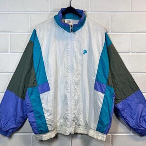 Pode incluir: Uma jaqueta corta-vento branca, azul, roxa e verde com fecho de correr e um pequeno logotipo "P" azul no peito.