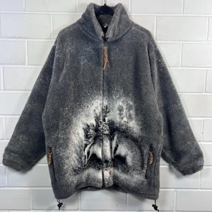 Könnte beinhalten: Graue Fleecejacke mit hohem Kragen und durchgehendem Reißverschluss. Die Jacke hat ein Wald- und Wolf-Design in Weiß und Grau auf der Vorderseite. Sie hat zwei Seitentaschen und einen Kordelzug am unteren Saum.
