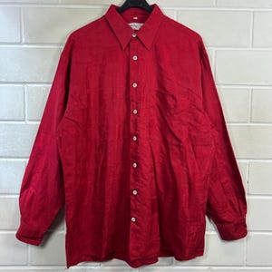 Könnte beinhalten: Ein langärmeliges rotes Button-Down-Hemd mit klassischem Kragen. Das Hemd hat ein strukturiertes Muster und weiße Knöpfe. Das Kleidungsstück hängt an einem schwarzen Kleiderbügel vor einer weißen Backsteinmauer.