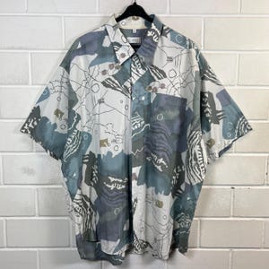 Könnte beinhalten: Kurzärmeliges Button-Down-Hemd mit einem abstrakten Muster in Weiß-, Blau-, Grau- und Brauntönen. Das Hemd hat einen Kragen, eine Brusttasche und eine lockere Passform. Der Stoff scheint leicht zu sein.