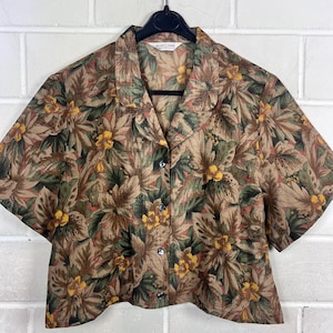 Peut inclure: Chemise à manches courtes avec un motif floral dans les tons marron, vert et jaune. La chemise est boutonnée et suspendue à un cintre noir. L'étiquette indique "MARILYNNE". Fond de mur en briques blanches.