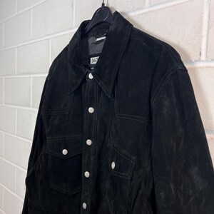 Peut inclure: Veste en daim noire avec une fermeture boutonn&eacute;e, deux poches de poitrine et un col classique. La veste est suspendue &agrave; un cintre noir contre un mur de briques blanches. Les boutons sont argent&eacute;s.