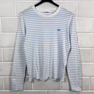 May include: A white long-sleeved shirt with horizontal blue stripes. It has a crew neck and a small blue Levi's logo on the chest. The shirt is displayed on a black hanger against a white brick wall.