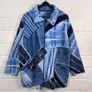 Könnte beinhalten: Blaue Fleecejacke mit Patchwork-Design. Die Jacke hat eine Vielzahl von Blautönen und Mustern, darunter geometrische und gestreifte Designs. Sie hat einen durchgehenden Reißverschluss, zwei Taschen und einen Kragen.