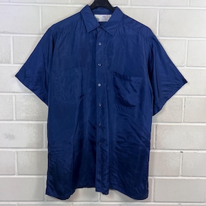 以下が含まれることがあります： ネイビーブルーの半袖シャツ。ボタンダウンで、胸に2つのポケットが付いています。軽くて少し光沢のある生地で作られています。黒いハンガーに掛けられ、白いレンガの壁を背景にしています。