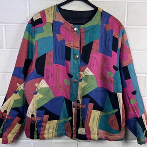 Könnte beinhalten: Bunte Jacke mit Knopfleiste und geometrischem Muster in Pink, Blau, Grün, Gelb und Schwarz. Die Jacke hat einen runden Kragen und goldfarbene Knöpfe. Sie hängt an einem schwarzen Kleiderbügel.