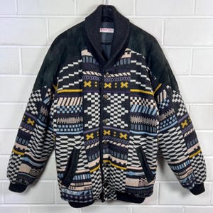 Puede incluir: Chaqueta vintage con un patrón geométrico en negro, blanco, amarillo y azul. La chaqueta tiene un cuello y detalles de ante negro, con cierre de botones y bolsillos. La marca "Terno" es visible en el cuello.