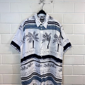 Puede incluir: Camisa blanca de manga corta con botones, con un estampado de palmeras y rayas. La camisa tiene una etiqueta de John Gee.