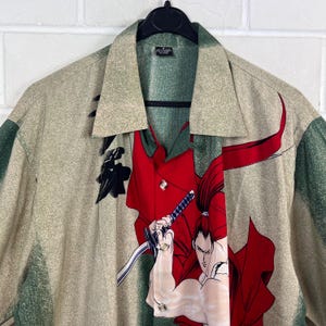 K&ouml;nnte beinhalten: Ein Hemd mit Kn&ouml;pfen mit beigem und gr&uuml;nem Muster. Das Hemd hat eine rote Tafel mit einer Grafik eines Samurai, der ein Schwert h&auml;lt. Kragen und Manschetten sind gr&uuml;n.