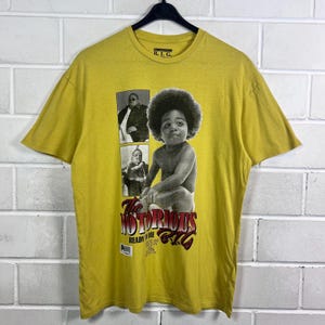 Könnte beinhalten: Senfgelbes T-Shirt mit einer Schwarz-Weiß-Grafik von The Notorious B.I.G. als Baby, mit den Worten "The Notorious B.I.G." in Rot und Weiß. Das Shirt hat auch zwei kleinere Bilder des Rappers.