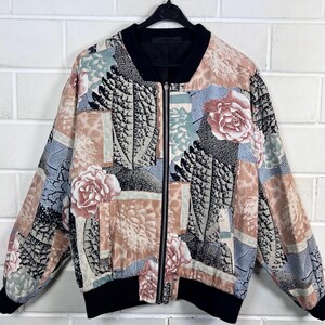 Op de afbeelding: Een bomberjack met bloemenprint en een zwarte kraag, manchetten en zoom. De jas heeft een ritssluiting over de hele lengte en een kleurrijk patroon van bloemen en abstracte vormen in roze, blauw en beige.