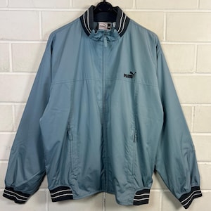 May include: A teal Puma track jacket with a full zip. The jacket has a black collar and cuffs with white stripes. The Puma logo is embroidered in black on the chest. Two zipped pockets are visible.