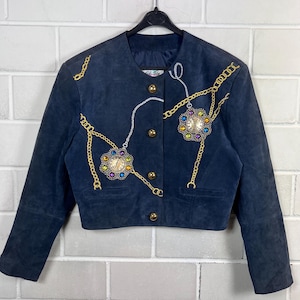 Peut inclure: Veste en daim bleu marine ornée de chaînes et de bijoux dorés. La veste courte est dotée de boutons dorés et d'un col rond. Le design comprend deux pendentifs horloge sertis de bijoux et des accents de chaîne dorée.