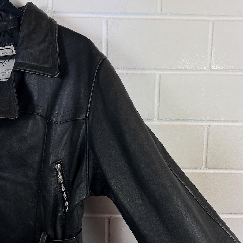 May include: Black leather jacket with a notched collar and a zipper pocket on the left sleeve. The jacket has a belt and a label on the collar. The jacket is made of a smooth, shiny leather material.