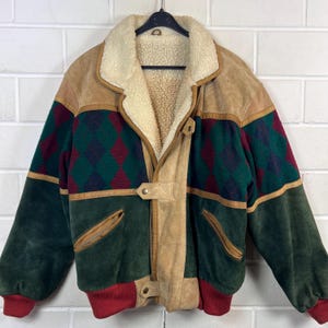 May include: A vintage suede jacket with a shearling collar. The jacket has a colour block design with green, tan, and burgundy accents. The sleeves are green, and the cuffs are red. The jacket is hanging on a black hanger.
