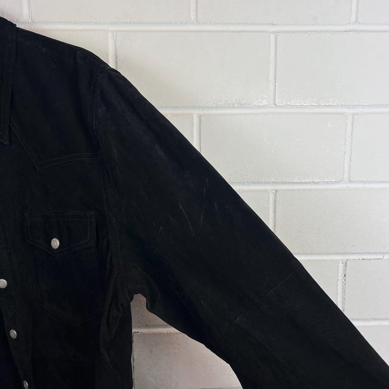 Peut inclure: Gros plan sur une veste en daim noire. La veste pr&eacute;sente un col classique, deux poches de poitrine avec des fermetures &agrave; boutons et des boutons argent&eacute;s sur le devant et aux poignets. Le tissu a un aspect textur&eacute;.