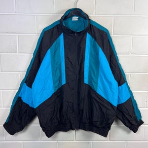 May include: A vintage track jacket in black, teal, and blue, with a full zip. The jacket has a colour block design with teal and blue vertical stripes on the front and sleeves. It hangs on a metal hanger against a white brick wall.