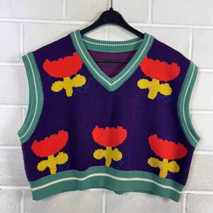 Op de afbeelding: Een paars gebreid vest met een V-hals en een groen-witte gestreepte bies. Het vest heeft een patroon van rode en gele bloemmotieven. Het vest is mouwloos en hangt aan een zwarte hanger tegen een witte bakstenen muur.