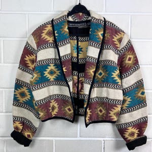 Op de afbeelding: Een kleurrijke jas met open voorkant en een geometrisch patroon in teal, bordeaux, geel en crème. De jas heeft zwarte biezen en manchetten. Het label luidt "NEW IDENTITY".