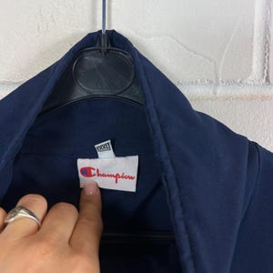 May include: Close-up of a navy blue Champion jacket. The tag features the Champion logo in red and blue, with the brand name in red script. The tag also indicates a size of XXL.