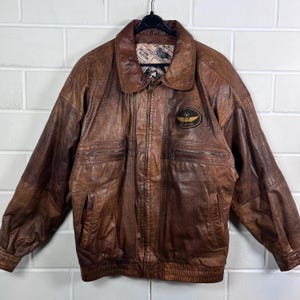 May include: A brown leather jacket with a zip-up front and a collared neckline. The jacket has two chest pockets and two lower pockets. An embroidered patch with an emblem is on the upper left chest. The jacket is hanging on a black hanger.