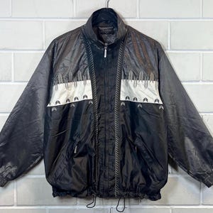 May include: Black and silver jacket with a zip fastening and a drawstring at the bottom. The jacket features a geometric pattern with white accents.