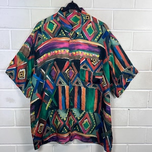 Könnte beinhalten: Ein kurzärmeliges Hemd mit Knopfleiste und einem lebendigen, mehrfarbigen, abstrakten geometrischen Muster. Das Design umfasst Rauten, Streifen und Pinselstrichmotive in Rosa, Grün, Blau, Orange und Gelb auf einem schwarzen Hintergrund.