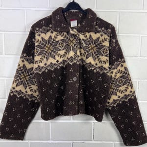 Può includere: Una giacca in pile marrone e color crema realizzata in materiale testurizzato e soffice. Il capo presenta un colletto, maniche lunghe e una chiusura frontale con bottoni. Un motivo ripetitivo ispirato ai fiocchi di neve adorna il petto.