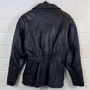May include: Black leather jacket with a notched collar, long sleeves, and a cinched waist. The jacket features a belt detail at the waist and a button closure. The back of the jacket has a yoke detail and vertical seams.