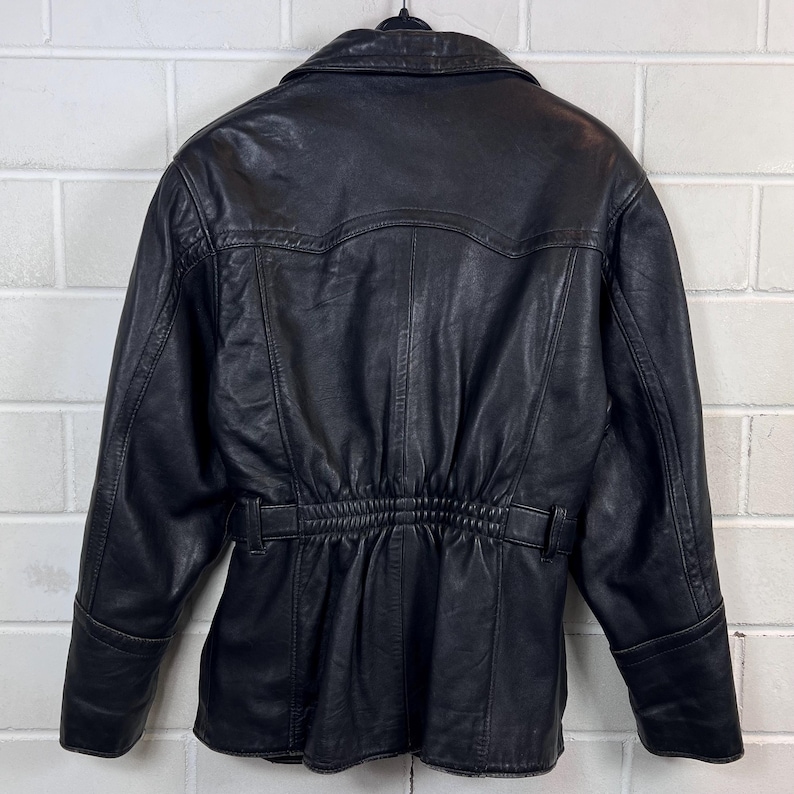 May include: Black leather jacket with a notched collar, long sleeves, and a cinched waist. The jacket features a belt detail at the waist and a button closure. The back of the jacket has a yoke detail and vertical seams.