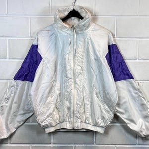 May include: A white windbreaker jacket with purple accents on the sleeves. The jacket has a zip closure and a drawstring waist.